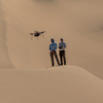 NASA Trials Mars Drones in Mojave Desert Two people stand atop a sand dune, facing the camera. They are both looking at a drone flying in front of them. The person on the right holds a controller. Other sand dunes behind them make up the background of the entire image.