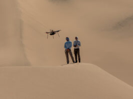 NASA Trials Mars Drones in Mojave Desert Two people stand atop a sand dune, facing the camera. They are both looking at a drone flying in front of them. The person on the right holds a controller. Other sand dunes behind them make up the background of the entire image.