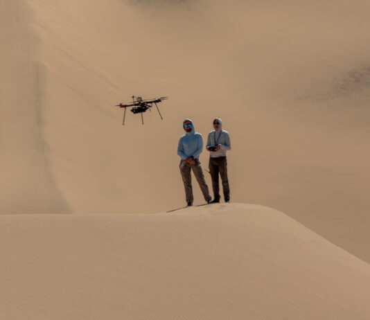 NASA Trials Mars Drones in Mojave Desert Two people stand atop a sand dune, facing the camera. They are both looking at a drone flying in front of them. The person on the right holds a controller. Other sand dunes behind them make up the background of the entire image.
