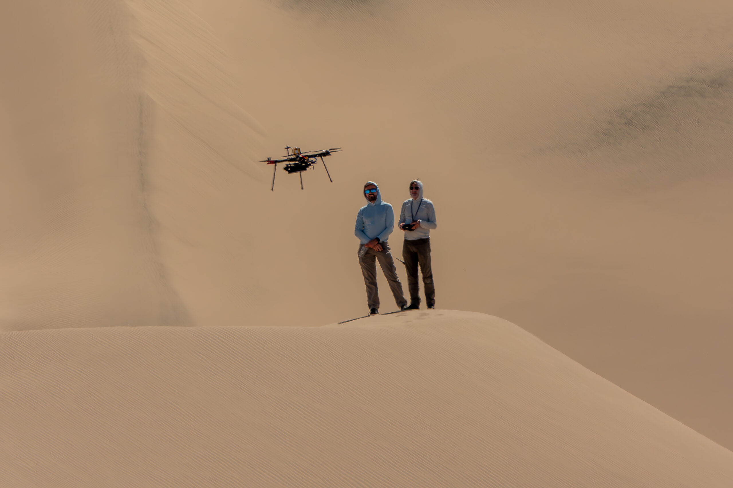 Two people stand atop a sand dune, facing the camera. They are both looking at a drone flying in front of them. The person on the right holds a controller. Other sand dunes behind them make up the background of the entire image.