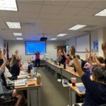 Virginia Classrooms Experience NASA Science Through STEM Educators’ Initiatives Approximately 25 people sitting at long tables raise their arms up in the air with their fingers tips spread out, waving arms from side to side.