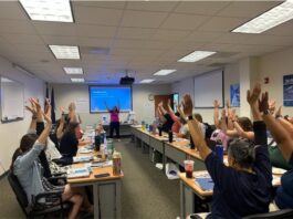 Virginia Classrooms Experience NASA Science Through STEM Educators’ Initiatives Approximately 25 people sitting at long tables raise their arms up in the air with their fingers tips spread out, waving arms from side to side.