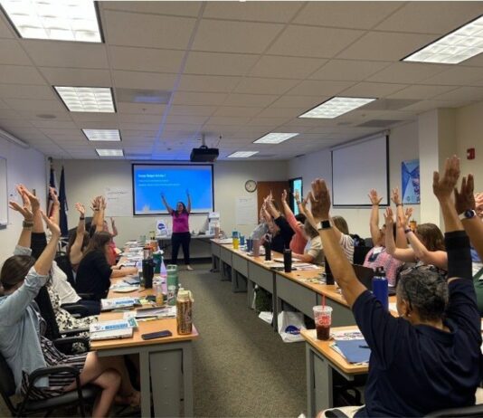 Virginia Classrooms Experience NASA Science Through STEM Educators’ Initiatives Approximately 25 people sitting at long tables raise their arms up in the air with their fingers tips spread out, waving arms from side to side.