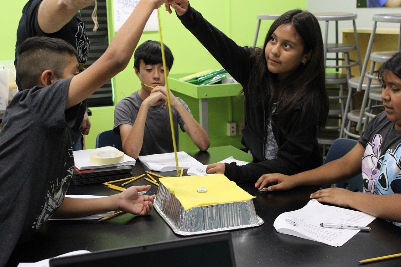 planets space hazards lauren shollenberger.jpg An out of school time educator holds a measuring tape as an elementary aged girl drops a weight to test the space craft shield her group created. Her group members, two boys and one girl, watch as she drops the weight.