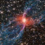 NASA Explores the Mysteries of the Red Spider Nebula A multicolored nebula stands out against the star-studded background of space. The nebula’s central star is hidden by a blotchy pinkish cloud of dust. A strong red light radiates from this area, lighting up the nearby dust. An elongated purple ‘S’ shape is centered on the heart of the nebula, while two blue lobes stretch diagonally outward to the edges of the image, making the entire structure look like a tilted hourglass.