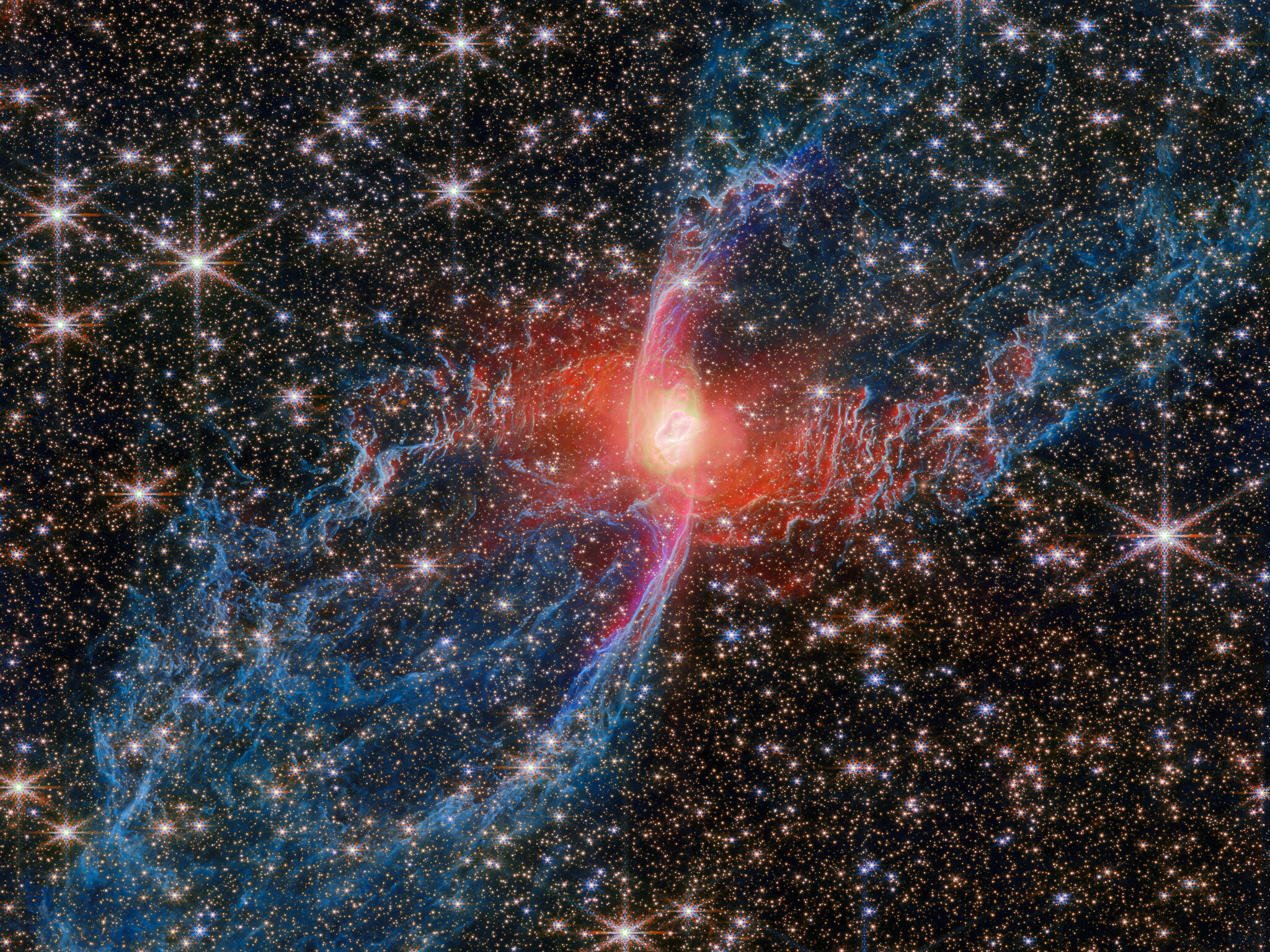 A multicolored nebula stands out against the star-studded background of space. The nebula’s central star is hidden by a blotchy pinkish cloud of dust. A strong red light radiates from this area, lighting up the nearby dust. An elongated purple ‘S’ shape is centered on the heart of the nebula, while two blue lobes stretch diagonally outward to the edges of the image, making the entire structure look like a tilted hourglass.