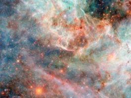 NASA’s Hubble Photographs Fluffy, Candy-Colored Clouds Part of a nebula in space. Layers of gas and dust clouds in different colors, from blue and green shades to pink, red, and black, each indicating light emitted by different molecules, comprise the nebula. The background cloud layers are thicker and puffier, though still translucent, and the upper layers are thin and bright at the edges. Behind the clouds are many small, mostly orange and some blue, stars.