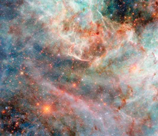 NASA’s Hubble Photographs Fluffy, Candy-Colored Clouds Part of a nebula in space. Layers of gas and dust clouds in different colors, from blue and green shades to pink, red, and black, each indicating light emitted by different molecules, comprise the nebula. The background cloud layers are thicker and puffier, though still translucent, and the upper layers are thin and bright at the edges. Behind the clouds are many small, mostly orange and some blue, stars.