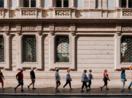 Explore Rome’s secret gems using an AI virtual guide A line of tourists, following a leader with a flag, on a city sidewalk with an ornate building behind them.