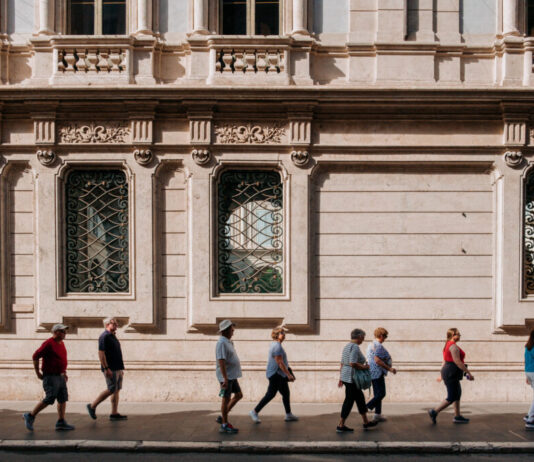 Explore Rome’s secret gems using an AI virtual guide A line of tourists, following a leader with a flag, on a city sidewalk with an ornate building behind them.