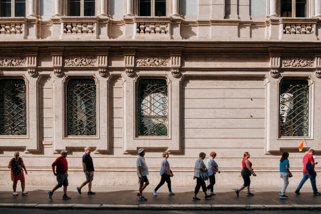 rometouristvirtualassistant 6 1024x683.jpg A line of tourists, following a leader with a flag, on a city sidewalk with an ornate building behind them.