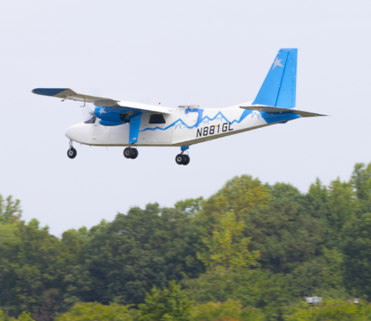 NASA Trials Lightweight Antennas for Future Airspace Advancements A white-and-blue Britten-Norman Defender aircraft outfitted for a flight test with an advanced phased array antenna prototype. The array is mounted on top of the fuselage near the center of the airplane. The plane is seen in flight over trees.