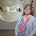Childhood Telescope Ignites Christina Zeringue’s NASA Career Christina Zeringue wearing a gray cardigan stands before a silver NASA insignia