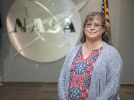 Childhood Telescope Ignites Christina Zeringue’s NASA Career Christina Zeringue wearing a gray cardigan stands before a silver NASA insignia