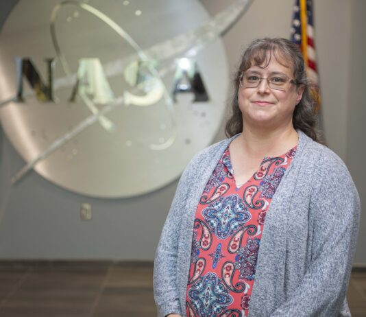 Childhood Telescope Ignites Christina Zeringue’s NASA Career Christina Zeringue wearing a gray cardigan stands before a silver NASA insignia