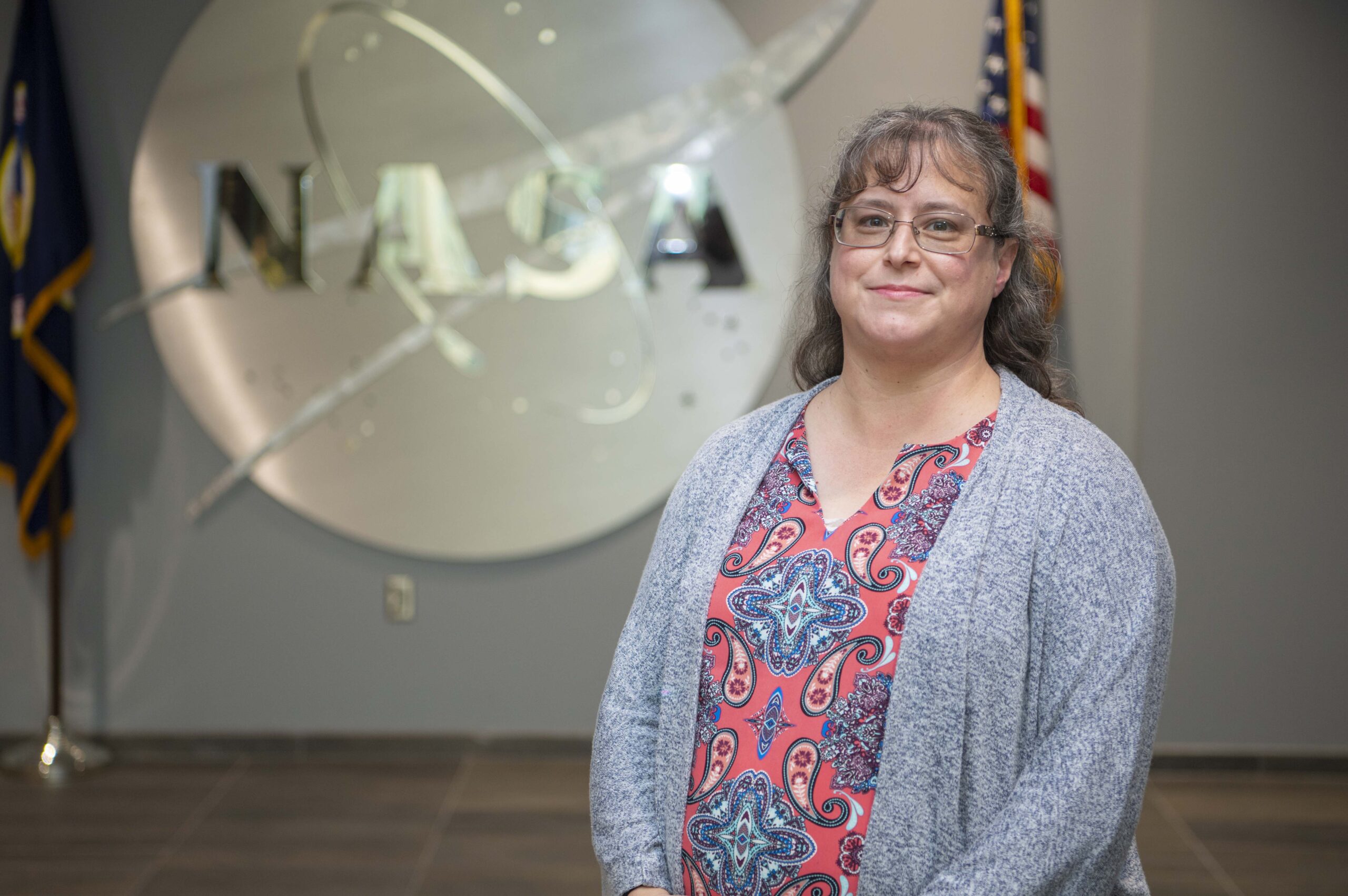s25 016 christina zeringue.jpg Christina Zeringue wearing a gray cardigan stands before a silver NASA insignia