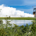 NASA Conducts Trial of Innovative RS-25 Rocket Engine A row of bushes frame the photo as vapor clouds seen in the distance erupt towards the sky during an engine test on the Fred Haise Test Stand at NASA Stennis