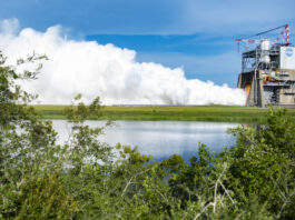 NASA Conducts Trial of Innovative RS-25 Rocket Engine A row of bushes frame the photo as vapor clouds seen in the distance erupt towards the sky during an engine test on the Fred Haise Test Stand at NASA Stennis