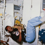 NASA’s Space Mission Features Saxophone Performance A Black man plays a saxophone while floating. He wears a black short sleeve shirt with patches on it, blue pants, and gray shoes. He is inside the space shuttle Challenger.