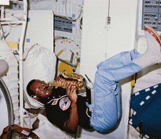 NASA’s Space Mission Features Saxophone Performance A Black man plays a saxophone while floating. He wears a black short sleeve shirt with patches on it, blue pants, and gray shoes. He is inside the space shuttle Challenger.