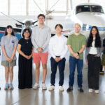 NASA’s 2025 SARP East Atmospheric Chemistry Group Announced A group of eight people stand together inside a hangar with a somewhat shiny floor. In the background is a small white plane with a blue stripe, and large windows behind that.