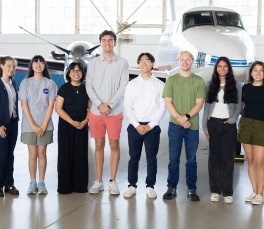 NASA’s 2025 SARP East Atmospheric Chemistry Group Announced A group of eight people stand together inside a hangar with a somewhat shiny floor. In the background is a small white plane with a blue stripe, and large windows behind that.