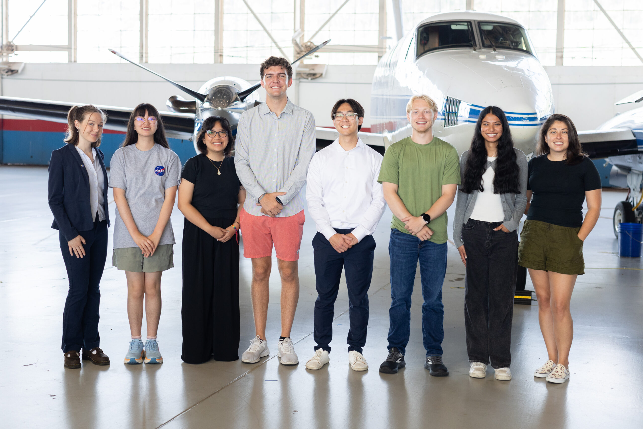 sarp east 6234.jpg A group of eight people stand together inside a hangar with a somewhat shiny floor. In the background is a small white plane with a blue stripe, and large windows behind that.