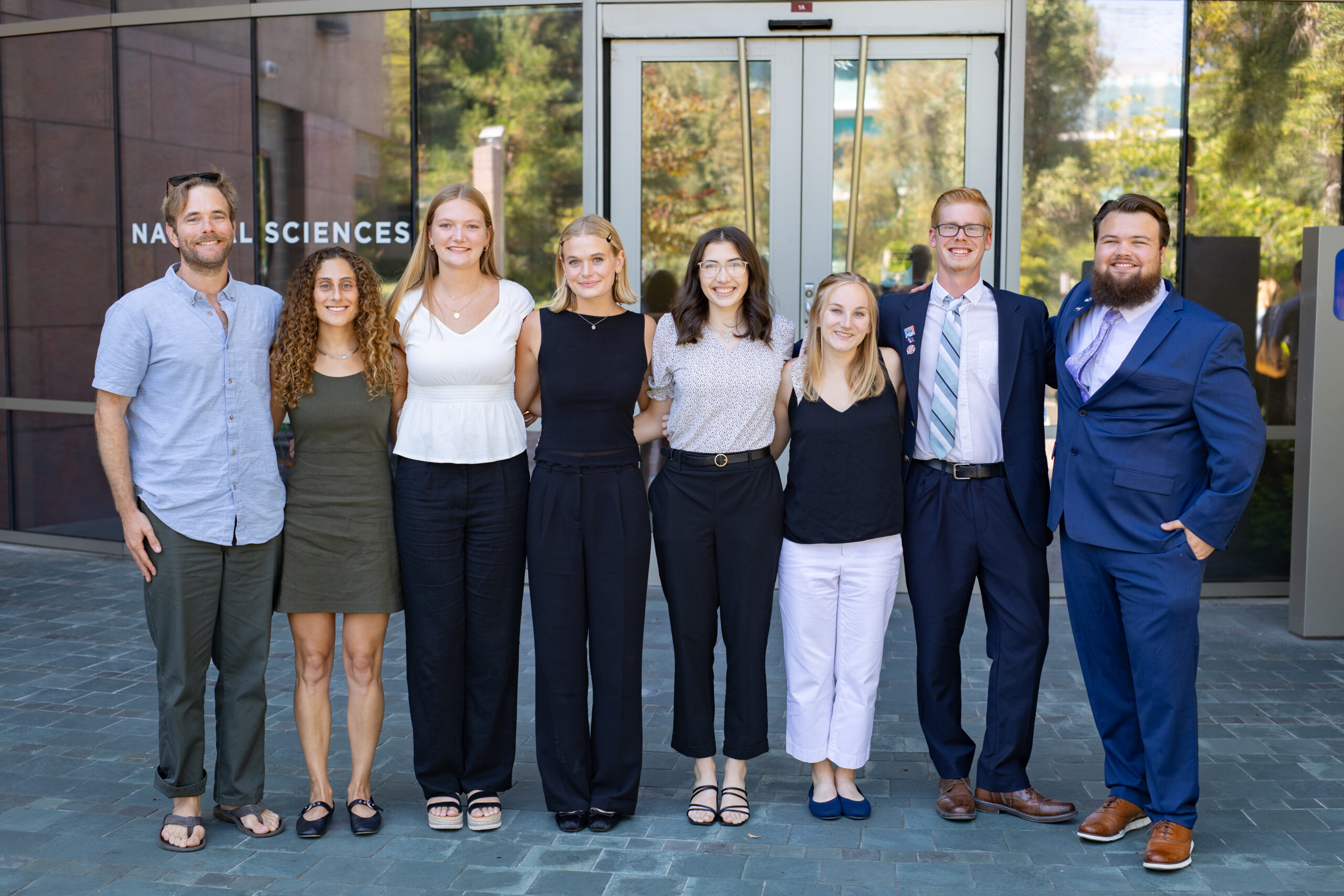 sarpwest2024 loiacono 1613.jpg A group of eight people. the first a professor and the other seven college age, stand in a line in professional attire. Behind them is a glass building with glass doors, reflecting green trees.