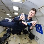 NASA Interns Explore Aerospace Research in Microgravity Conditions A young man in a navy blue flight suit floats inside a microgravity simulation aircraft, smiling as he demonstrates a Wound Irrigation System using two syringes and a small device. The padded interior of the aircraft is visible behind him, along with logos for Zero-G and NASA SEES on the wall. He appears weightless, mid-air, during a parabolic flight aboard the ZERO-G G-FORCE ONE®.