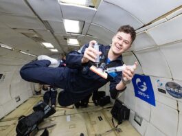 NASA Interns Explore Aerospace Research in Microgravity Conditions A young man in a navy blue flight suit floats inside a microgravity simulation aircraft, smiling as he demonstrates a Wound Irrigation System using two syringes and a small device. The padded interior of the aircraft is visible behind him, along with logos for Zero-G and NASA SEES on the wall. He appears weightless, mid-air, during a parabolic flight aboard the ZERO-G G-FORCE ONE®.