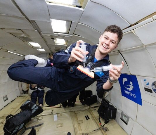 NASA Interns Explore Aerospace Research in Microgravity Conditions A young man in a navy blue flight suit floats inside a microgravity simulation aircraft, smiling as he demonstrates a Wound Irrigation System using two syringes and a small device. The padded interior of the aircraft is visible behind him, along with logos for Zero-G and NASA SEES on the wall. He appears weightless, mid-air, during a parabolic flight aboard the ZERO-G G-FORCE ONE®.