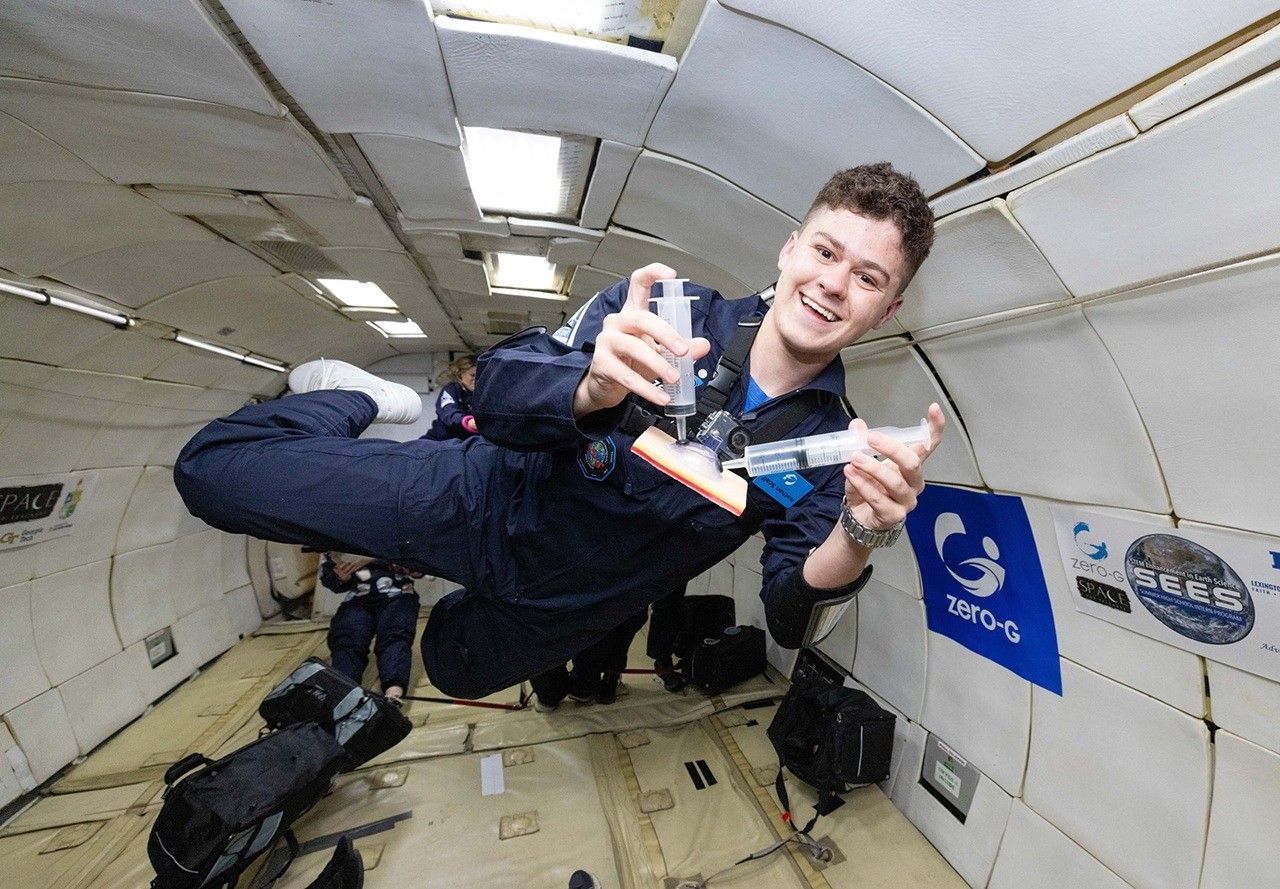sees student nathan scalf laura tomlin.jpg A young man in a navy blue flight suit floats inside a microgravity simulation aircraft, smiling as he demonstrates a Wound Irrigation System using two syringes and a small device. The padded interior of the aircraft is visible behind him, along with logos for Zero-G and NASA SEES on the wall. He appears weightless, mid-air, during a parabolic flight aboard the ZERO-G G-FORCE ONEĀ®.