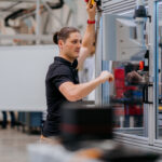 Siemens, thyssenkrupp leverage AI to address manufacturing skill shortages A man with a ponytail in a dark blue polo shirt working on the exterior of a machine in a glass box.