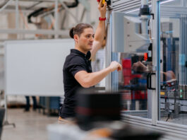 Siemens, thyssenkrupp leverage AI to address manufacturing skill shortages A man with a ponytail in a dark blue polo shirt working on the exterior of a machine in a glass box.