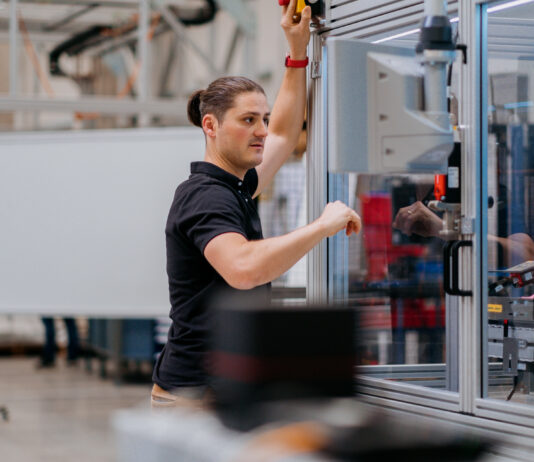 Siemens, thyssenkrupp leverage AI to address manufacturing skill shortages A man with a ponytail in a dark blue polo shirt working on the exterior of a machine in a glass box.