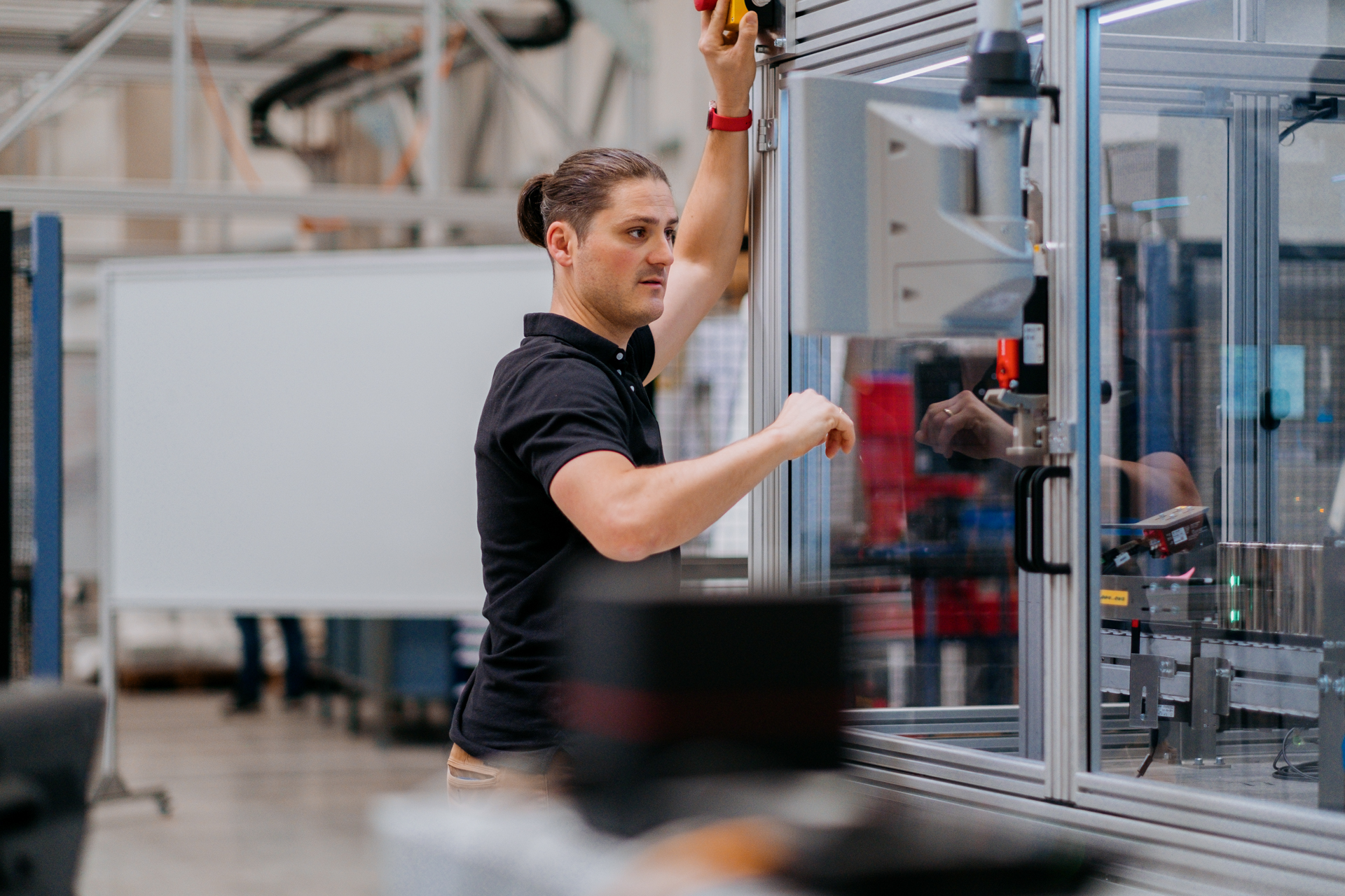 siemens industrial copilot 01.jpg.jpg A man with a ponytail in a dark blue polo shirt working on the exterior of a machine in a glass box.