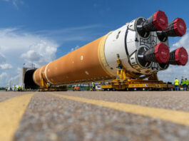 NASA’s Marshall Star – July 17, 2024 Edition Released The core stage of the Artemis Space Launch System being loaded on a covered barge. The stage is a large cylinder shape with the engines facing toward the camera on two yellow transporters that are guiding the stage into a covered grey container in the background. The body of the cylinder is mostly an orange color and white around the bottom. The four engines on the bottom are covered with red material.