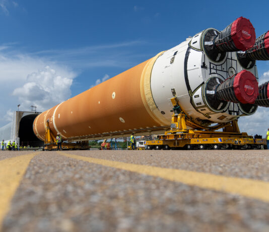 NASA’s Marshall Star – July 17, 2024 Edition Released The core stage of the Artemis Space Launch System being loaded on a covered barge. The stage is a large cylinder shape with the engines facing toward the camera on two yellow transporters that are guiding the stage into a covered grey container in the background. The body of the cylinder is mostly an orange color and white around the bottom. The four engines on the bottom are covered with red material.