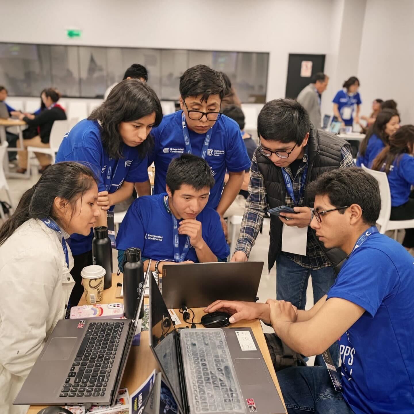 Six people look at a laptop computer in a classroom setting. Four wear blue t-shirts, one wears a white jacket, and one wears a plaid shirt with a black vest.