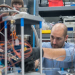 NASA Evaluates Innovative Fuel for Deep Space Missions A man wearing a blue shirt leans over a tabletop to make adjustments to a Stirling engine testbed. The mechanism showcases three pillars, about 12 inches in height with a metal cover. Orange winding cords are visible.