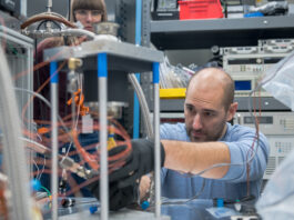 NASA Evaluates Innovative Fuel for Deep Space Missions A man wearing a blue shirt leans over a tabletop to make adjustments to a Stirling engine testbed. The mechanism showcases three pillars, about 12 inches in height with a metal cover. Orange winding cords are visible.