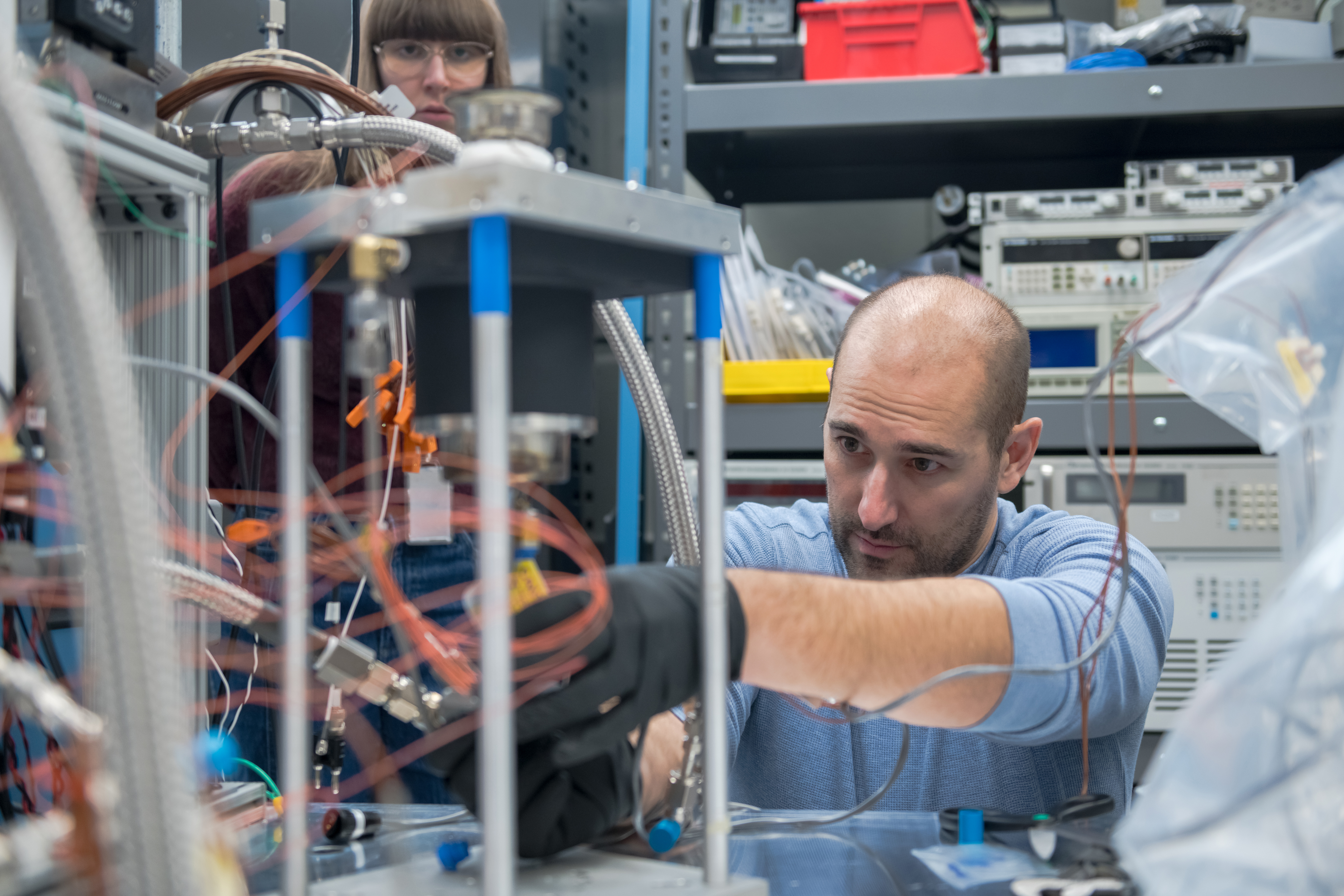 stirling additional image 1.jpg A man wearing a blue shirt leans over a tabletop to make adjustments to a Stirling engine testbed. The mechanism showcases three pillars, about 12 inches in height with a metal cover. Orange winding cords are visible.