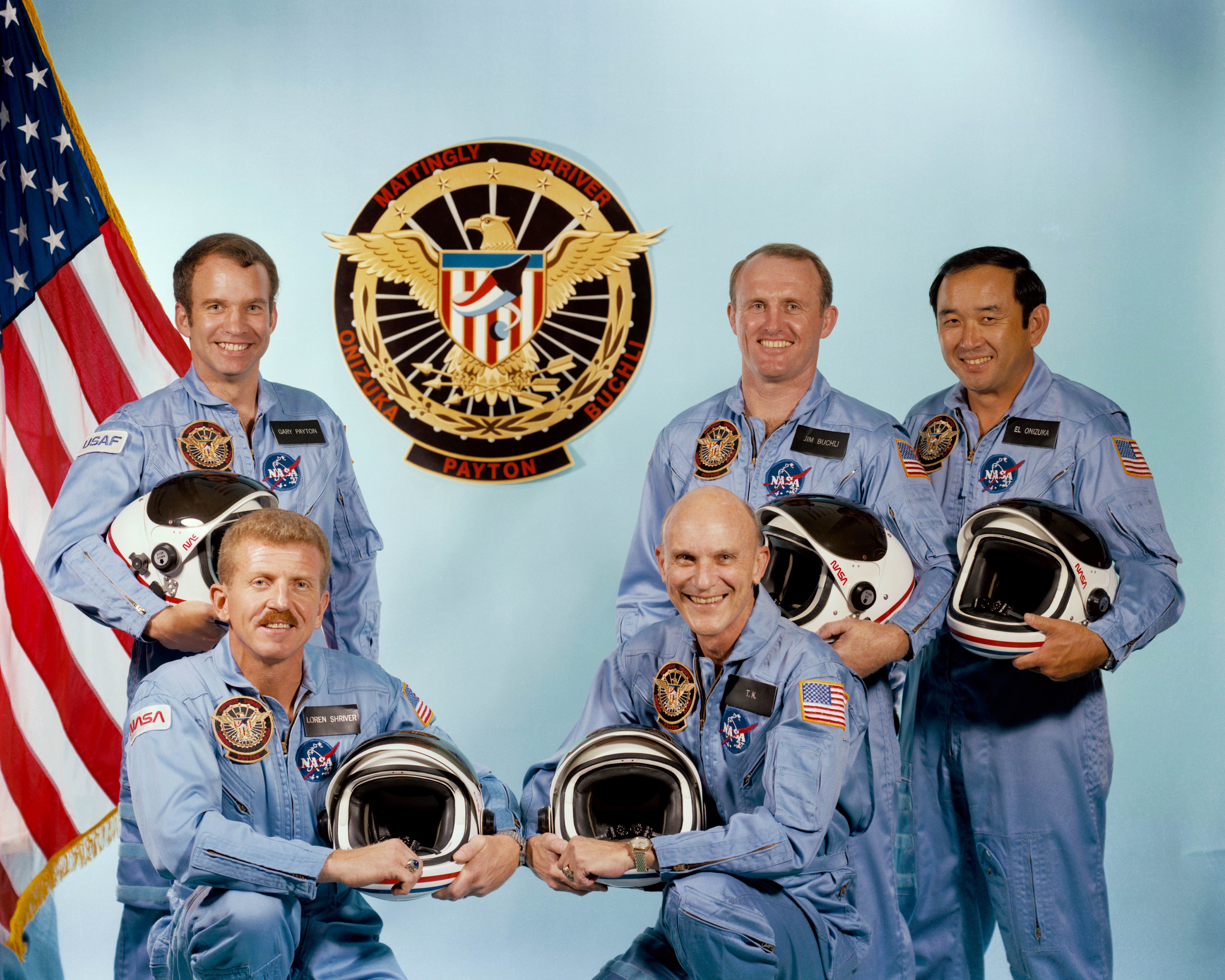 A group photo of five men clad in light blue flight suits, each holding a black and white helmet, with a US flag partially visible at left background and a round patch with a golden eagle in the background.