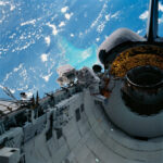 NASA’s Caribbean Adventure: A New Spacewalk Experience Two astronauts in white spacesuits inspect equipment outside of space shuttle Discovery during a spacewalk. The Caribbean Sea and part of the Bahama Islands chain is visible behind them.