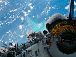 NASA’s Caribbean Adventure: A New Spacewalk Experience Two astronauts in white spacesuits inspect equipment outside of space shuttle Discovery during a spacewalk. The Caribbean Sea and part of the Bahama Islands chain is visible behind them.
