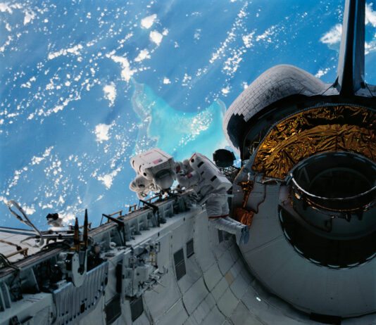 NASA’s Caribbean Adventure: A New Spacewalk Experience Two astronauts in white spacesuits inspect equipment outside of space shuttle Discovery during a spacewalk. The Caribbean Sea and part of the Bahama Islands chain is visible behind them.