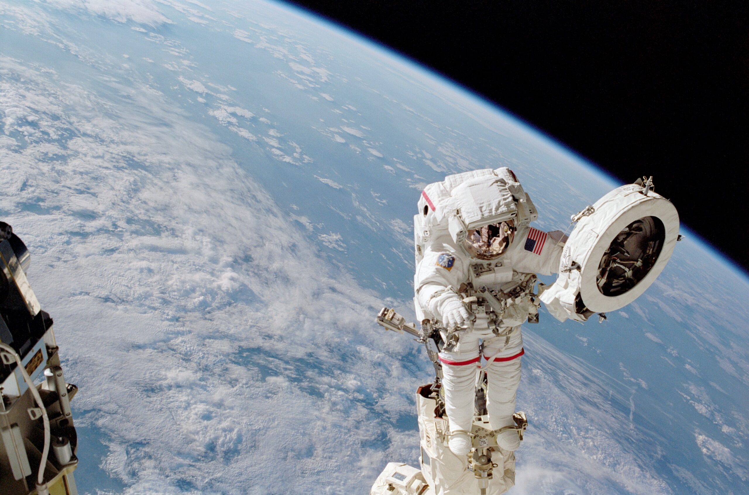 sts111 306 012orig.jpg An astronaut in a white spacesuit works on a fixture while outside of the International Space Station. He holds a large white circular object in his left hand. Earth can be seen behind him: the planet