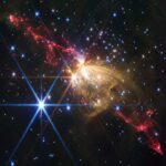 Webb Telescope Spots Massive Stellar Jet at Milky Way’s Edge Gaseous yellow-orange filaments look like a rose seen from the side and tilted slightly from upper left to lower right, slightly higher than the center of the frame. Extending from the rose to upper left and lower right are gaseous outflows that appear as red lobes that have an overall shape of tall, narrow triangles with rounded tips. Each red triangle is made up of wavy, irregular lines. Dozens of stars are scattered across the field. One particularly bright white star with eight diffraction spikes is located at the top of the yellow rose. Another bright blue star with even more prominent diffraction spikes is to its lower left. The background of space is black.