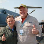 NASA’s Bob Swap Fosters Connections Through Science Diplomacy Woman wearing a green jacket and man wearing a tan button down shirt with the Goddard logo stand on a runway in front of a plane.