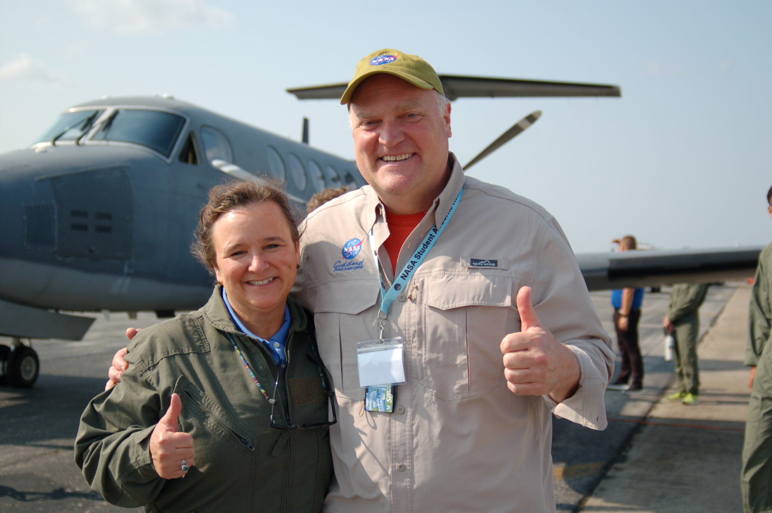 swap ksg close up sarp 2023.jpg Woman wearing a green jacket and man wearing a tan button down shirt with the Goddard logo stand on a runway in front of a plane.